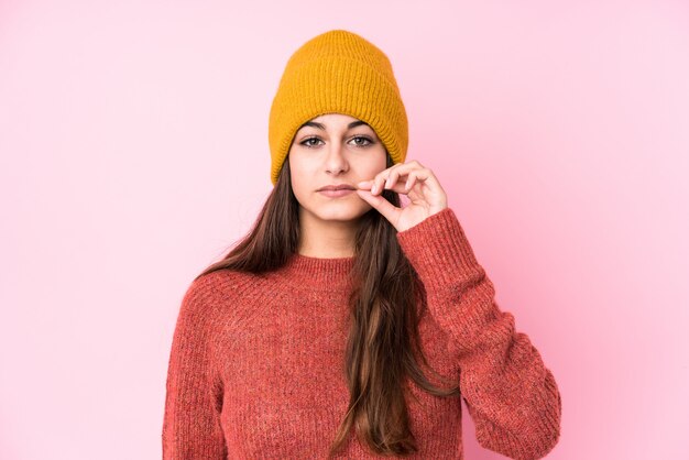 Giovane donna caucasica che indossa un berretto di lana con le dita sulle labbra mantenendo un segreto.