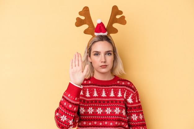 Giovane donna caucasica che indossa un cappello da renna di Natale isolato su sfondo giallo in piedi con la mano tesa che mostra il segnale di stop, impedendoti.