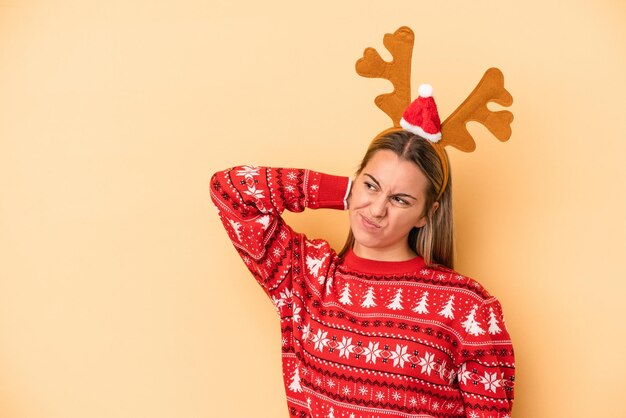 Giovane donna caucasica che indossa un cappello di renne di Natale isolato su sfondo giallo toccando la parte posteriore della testa, pensando e facendo una scelta.