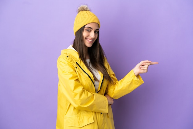 Giovane donna caucasica che indossa un cappotto antipioggia isolato su sfondo viola che punta il dito a lato