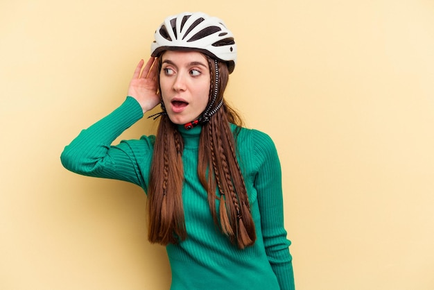 Giovane donna caucasica che indossa una bici da casco isolata su sfondo giallo cercando di ascoltare un pettegolezzo.
