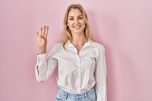 Giovane donna caucasica che indossa una camicia bianca casual su sfondo rosa che mostra e punta verso l'alto con le dita numero quattro mentre sorride fiducioso e felice.