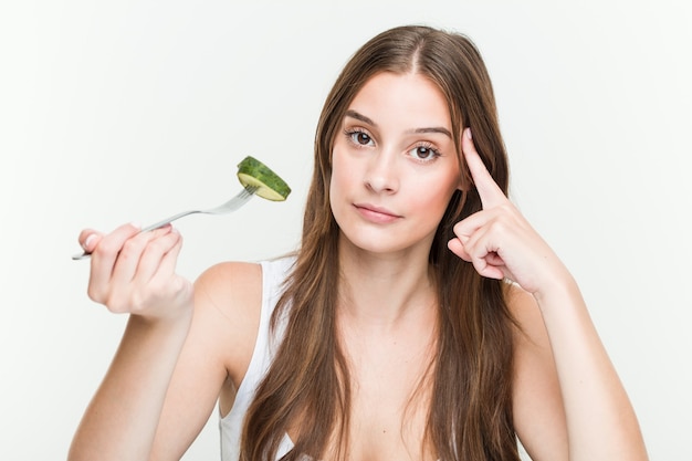 Giovane donna caucasica che mangia cetriolo che punta la tempia con il dito, pensando, concentrato su un compito.