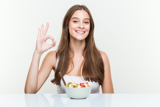 Giovane donna caucasica che mangia la ciotola di frutta allegra e sicura che mostra gesto giusto.