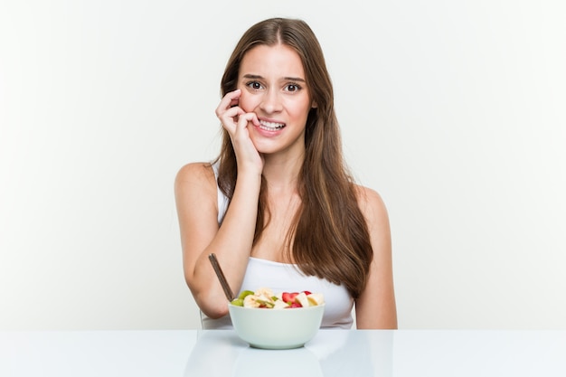 Giovane donna caucasica che mangia le unghie mordaci della ciotola di frutta, nervose e molto ansiose.