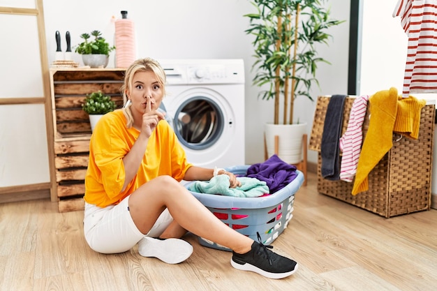 Giovane donna caucasica che mette i panni sporchi nella lavatrice chiedendo di stare zitta con il dito sulle labbra silenzio e concetto segreto