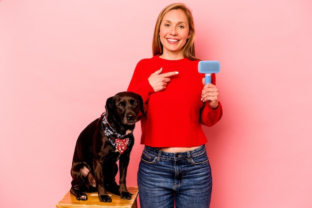 Giovane donna caucasica che pettina il cane isolato su sfondo rosa sorridente e indicando da parte mostrando qualcosa in uno spazio vuoto