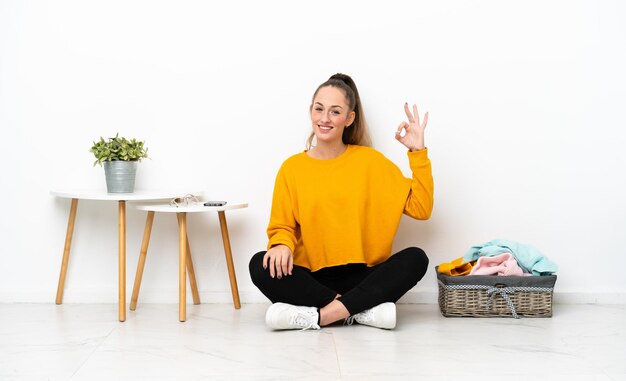 Giovane donna caucasica che piega i vestiti che si siede sul pavimento isolato su fondo bianco che mostra il segno giusto con le dita