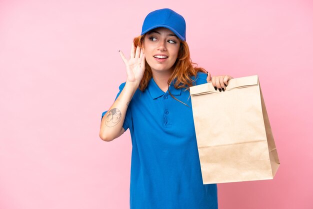 Giovane donna caucasica che prende un sacchetto di cibo da asporto isolato su sfondo rosa ascoltando qualcosa mettendo la mano sull'orecchio