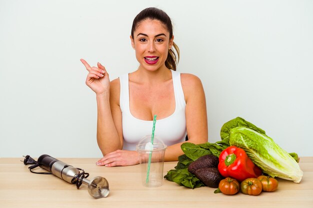 Giovane donna caucasica che prepara un frullato sano con le verdure sorridendo allegramente indicando con l'indice di distanza.