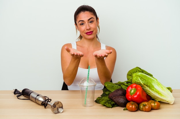 Giovane donna caucasica che prepara un frullato sano con verdure tenendo qualcosa con le palme, offrendo alla telecamera.