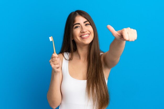 Giovane donna caucasica che pulisce i denti isolati su fondo blu che dà un pollice in su gesto