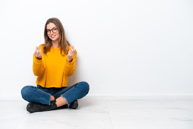 Giovane donna caucasica che si siede sul pavimento isolato su fondo bianco che fa gesto dei soldi