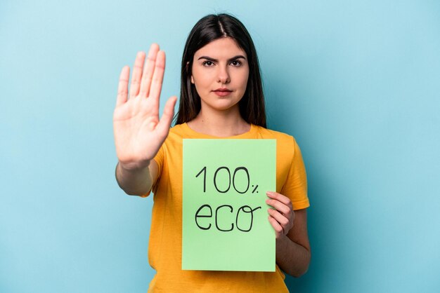 Giovane donna caucasica che tiene 100 eco cartello isolato su sfondo blu in piedi con la mano tesa che mostra il segnale di stop che ti impedisce