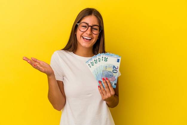 Giovane donna caucasica che tiene banconote isolate su sfondo giallo ricevendo una piacevole sorpresa, eccitata e alzando le mani.