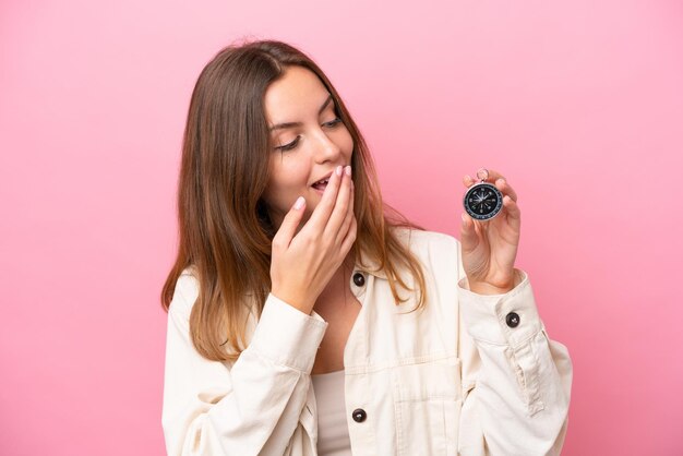 Giovane donna caucasica che tiene bussola isolata su sfondo rosa con espressione facciale sorpresa e scioccata