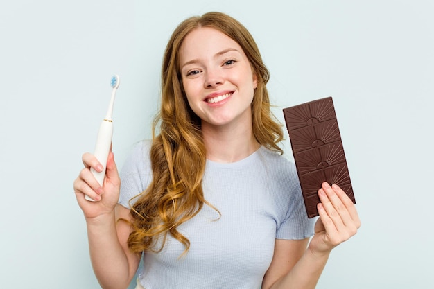 Giovane donna caucasica che tiene cioccolato e spazzolino da denti isolato su sfondo blu