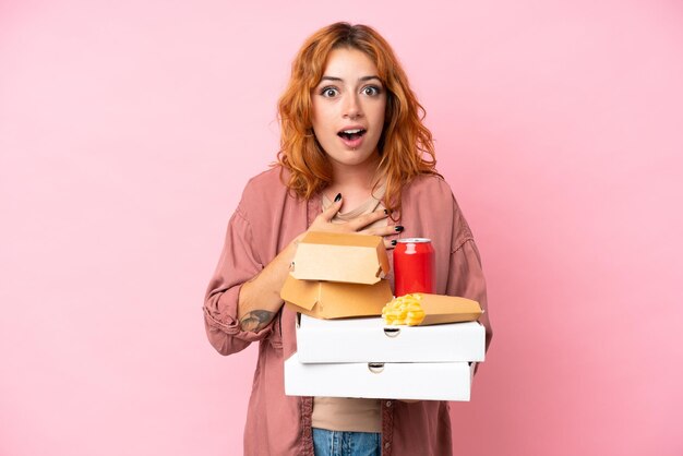 Giovane donna caucasica che tiene fast food isolato su sfondo rosa sorpreso e scioccato mentre guarda a destra