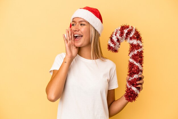 Giovane donna caucasica che tiene grande bastone di natale isolato su sfondo giallo gridando e tenendo il palmo vicino alla bocca aperta.