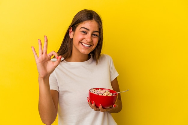 Giovane donna caucasica che tiene i cereali isolati su sfondo giallo allegro e fiducioso che mostra gesto ok.