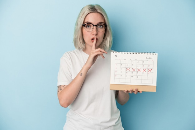 Giovane donna caucasica che tiene il calendario isolato su sfondo blu mantenendo un segreto o chiedendo silenzio.