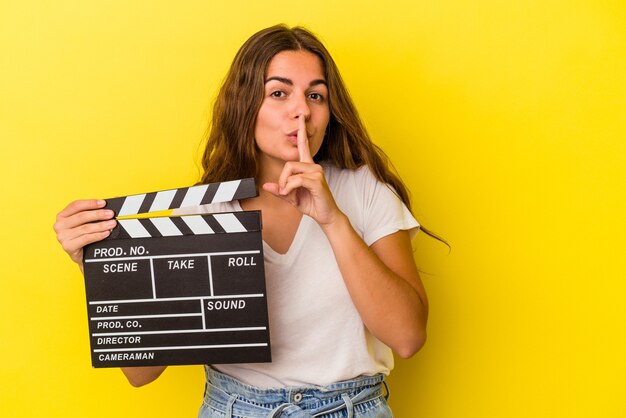 Giovane donna caucasica che tiene il ciak isolato su sfondo giallo mantenendo un segreto o chiedendo silenzio.