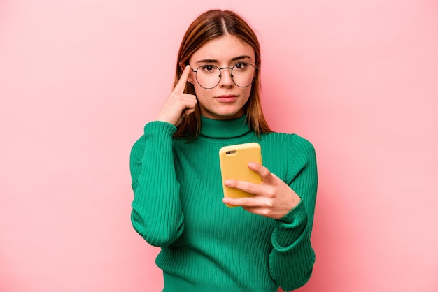 Giovane donna caucasica che tiene il telefono cellulare isolato su sfondo rosa che punta il tempio con il pensiero del dito concentrato su un compito