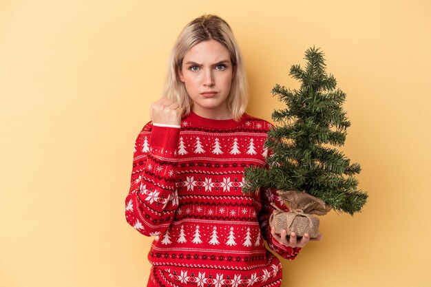 Giovane donna caucasica che tiene in mano un piccolo albero di natale isolato su sfondo giallo che mostra il pugno alla telecamera, espressione facciale aggressiva.