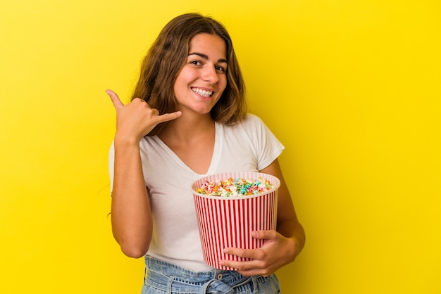 Giovane donna caucasica che tiene in mano un popcorn isolato su sfondo giallo che mostra un gesto di telefonata con le dita.