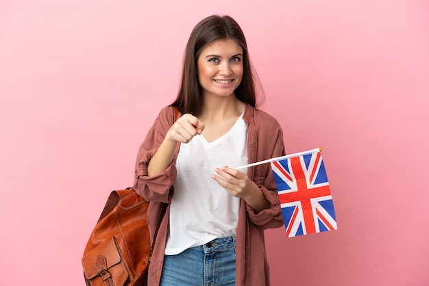 Giovane donna caucasica che tiene in mano una bandiera del Regno Unito isolata su sfondo rosa sorpresa e che punta davanti