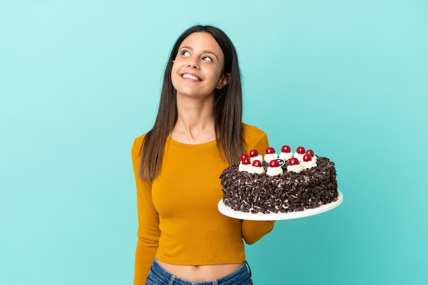 Giovane donna caucasica che tiene la torta di compleanno isolata su fondo blu che pensa ad un'idea mentre guarda in su