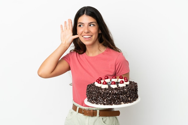 Giovane donna caucasica che tiene la torta di compleanno isolata su sfondo bianco ascoltando qualcosa mettendo la mano sull'orecchio