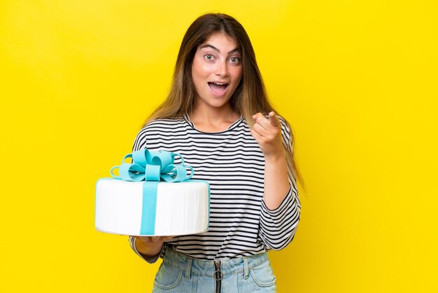 Giovane donna caucasica che tiene la torta di compleanno isolata su sfondo giallo sorpreso e puntato davanti