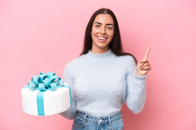 Giovane donna caucasica che tiene la torta di compleanno isolata su sfondo rosa che mostra e alza un dito in segno del meglio