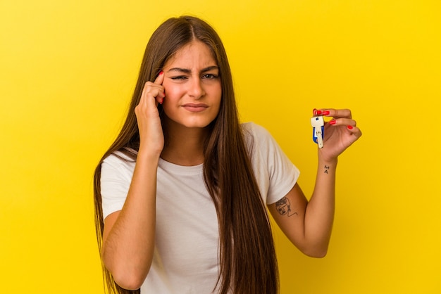 Giovane donna caucasica che tiene le chiavi di casa isolate su sfondo giallo che mostra un gesto di delusione con l'indice.