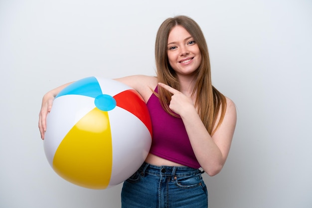 Giovane donna caucasica che tiene pallone da spiaggia isolato su sfondo bianco che punta al lato per presentare un prodotto