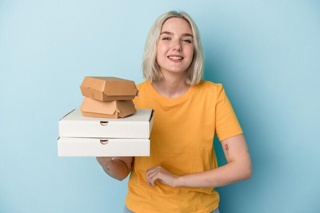 Giovane donna caucasica che tiene pizze e hamburger isolati su sfondo blu ridendo e divertendosi