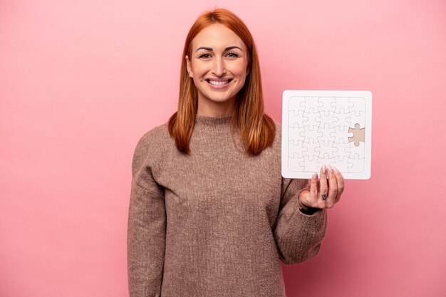 Giovane donna caucasica che tiene puzzle isolato su sfondo rosa felice, sorridente e allegro.
