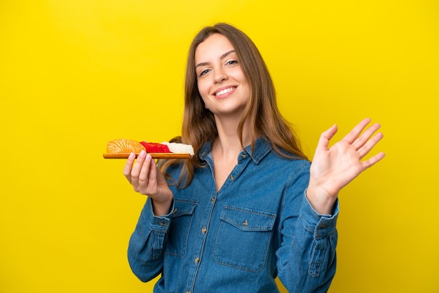 Giovane donna caucasica che tiene sashimi isolato su sfondo giallo salutando con la mano con un'espressione felice