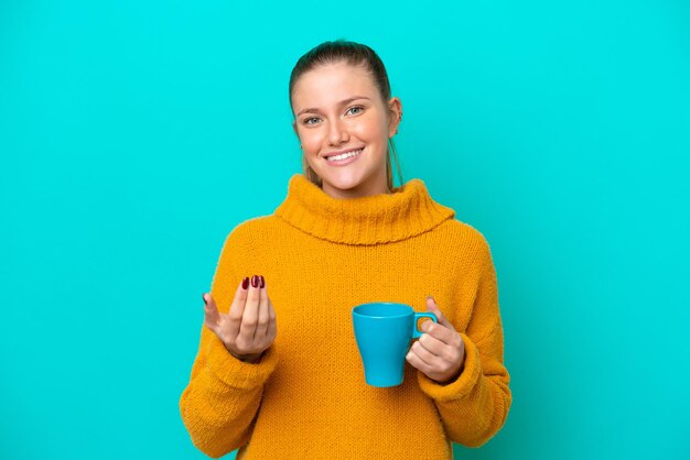 Giovane donna caucasica che tiene tazza isolata su sfondo blu che invita a venire con la mano Felice che tu sia venuto