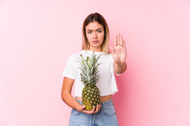 Giovane donna caucasica che tiene un ananas che sta con il fanale di arresto di rappresentazione della mano tesa, impedendovi.