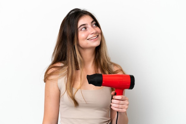 Giovane donna caucasica che tiene un asciugacapelli isolato su sfondo bianco alzando lo sguardo mentre sorride