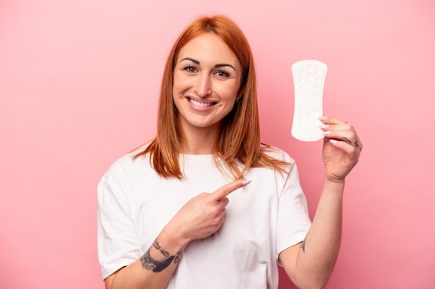 Giovane donna caucasica che tiene un assorbente igienico isolato su sfondo rosa sorridente e indicando da parte, mostrando qualcosa in uno spazio vuoto.