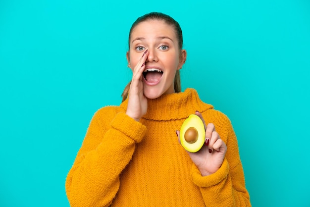 Giovane donna caucasica che tiene un avocado isolato su sfondo blu che grida con la bocca spalancata