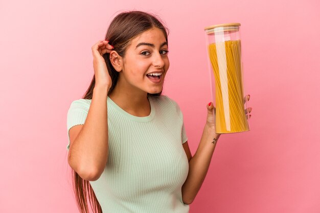 Giovane donna caucasica che tiene un barattolo di pasta isolato su sfondo rosa cercando di ascoltare un pettegolezzo.