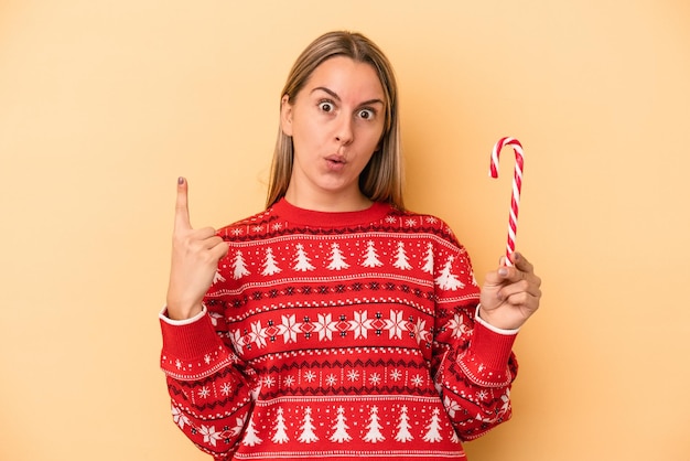 Giovane donna caucasica che tiene un bastone di natale isolato su sfondo giallo con qualche grande idea, concetto di creatività.