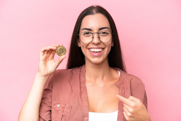 Giovane donna caucasica che tiene un Bitcoin isolato su sfondo rosa con espressione facciale a sorpresa