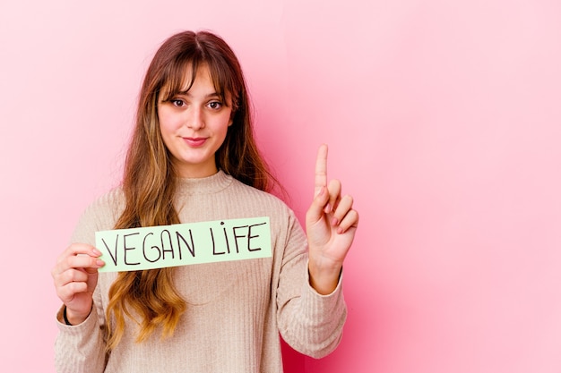 Giovane donna caucasica che tiene un cartello di vita vegano isolato che mostra il numero uno con il dito.
