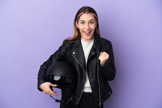 Giovane donna caucasica che tiene un casco del motociclo isolato su viola che celebra una vittoria nella posizione del vincitore
