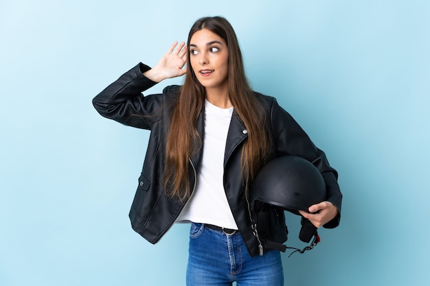 Giovane donna caucasica che tiene un casco del motociclo isolato sull'azzurro che ascolta qualcosa mettendo la mano sull'orecchio